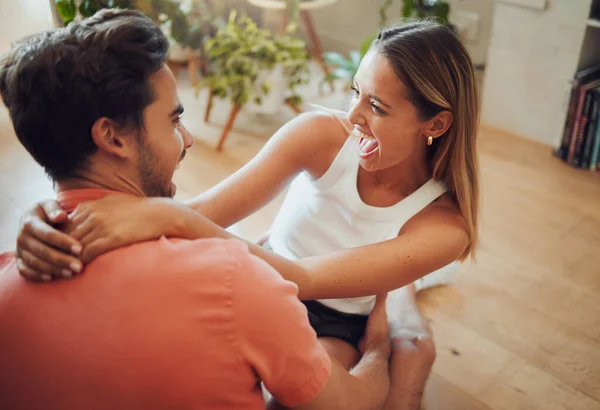 Funny young caucasian couple being playful laughing and having fun while sitting together at home. Young woman sitting on boyfriends lap putting her arms around him while they look at each other.