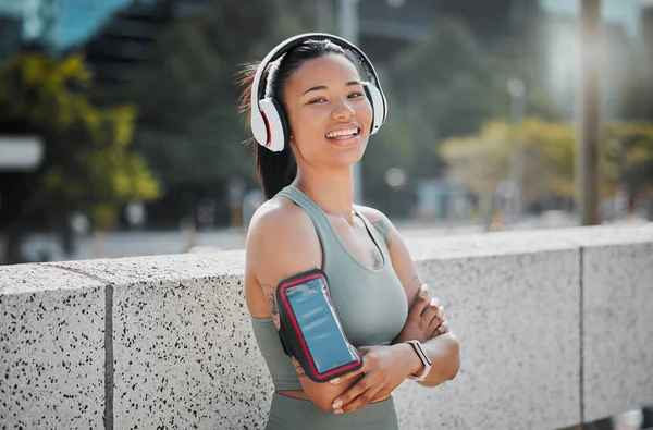 Young mixed race female athlete wearing headphones armband while listening to music and standing with her arms crossed exercising outside in the city. Positive, happy, workout.