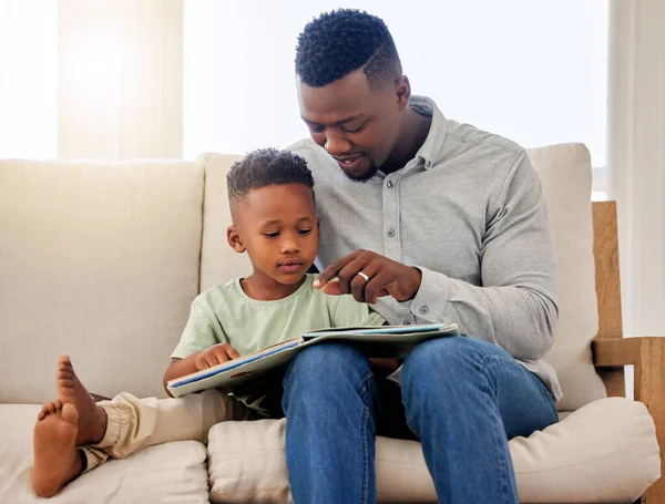 African american dad pointing at picture in storybook. Adorable little boy sitting with his father and reading a storybook together on the couch at home . Father and son spending time together.