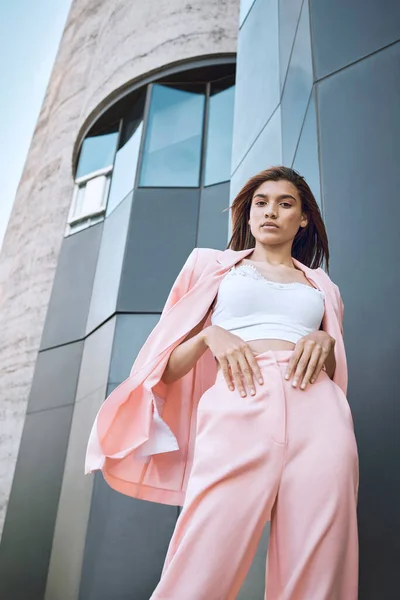 Portrait of a young trendy and confident mixed race woman looking stylish while posing and chilling time in the city. Fashionable hispanic woman wearing pink clothes and standing outside downtown.