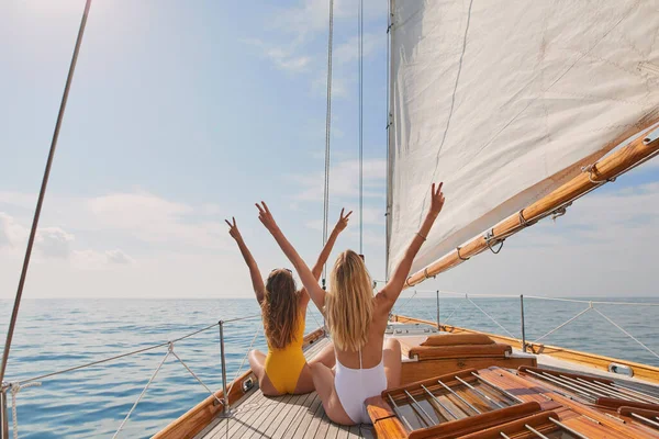 Two cheerful friends with arms raised making peace signs celebrating on boat cruise together. Two excited women in swimwear on holiday cruise celebrating cheering making peace signs in the air.