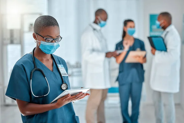 Young african american female doctor working on a digital tablet and wearing a mask while working at a hospital with colleagues. Black female nurse doing research on a digital tablet at work at a