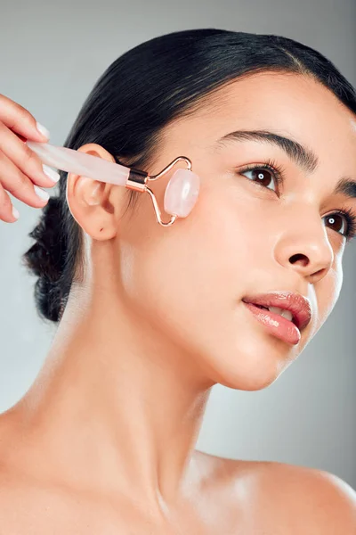 One beautiful mixed race woman using a rose quartz derma roller during a selfcare grooming routine. Young hispanic woman using anti ageing tool against grey copyspace background.