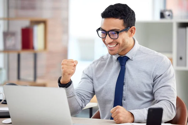 Young happy mixed race businessman cheering with his fist in joy while working on a laptop at work. Cheerful hispanic male businessperson celebrating success and victory in an office.