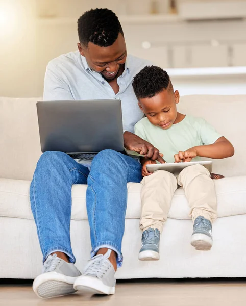 Happy cheerful young african american man using a laptop while helping his son with a digital tablet sitting on the couch at home. Father working and sending an email while bonding with his son.