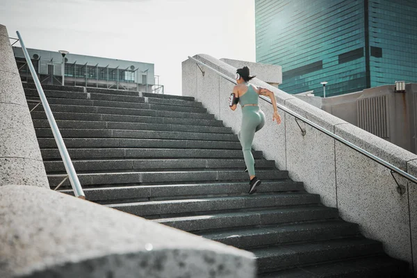 Rear view of a female wearing athlete wearing a phone armband and headphones while running up the steps of a building outside. Young female focused on her speed, body, fitness and cardio health while