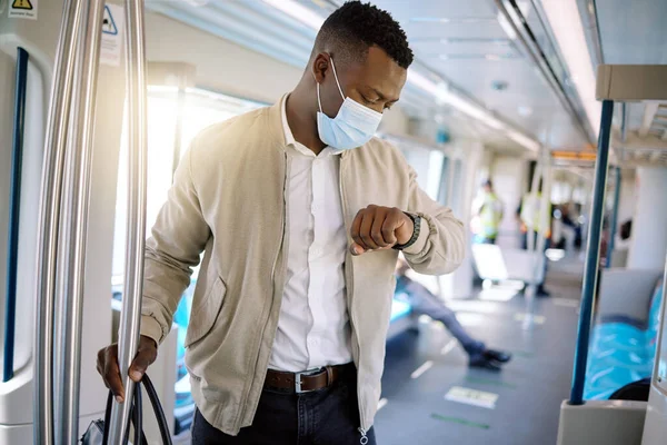 Black businessman travelling alone. A young african american businessman standing on a train while checking the time on her watch and wearing a mask during his commute to the city.