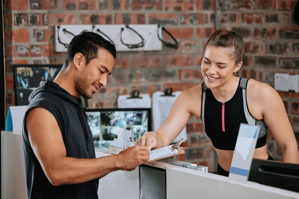 Asian man signing up for gym membership with caucasian trainer. Young coach behind reception pointing and showing where to sign. Two active and fit people standing together in health and fitness club.