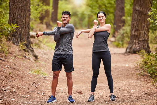 Portrait of a happy young male and female athlete stretching their arms before a run outside in nature. Two fit sportspeople doing warm-up exercises in pine forest on a sunny day.