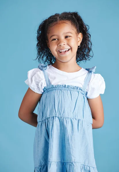 Happy smiling little girl standing with her hands behind her back against blue studio background. Cheerful mixed race kid in casual denim dress and white tshirt.