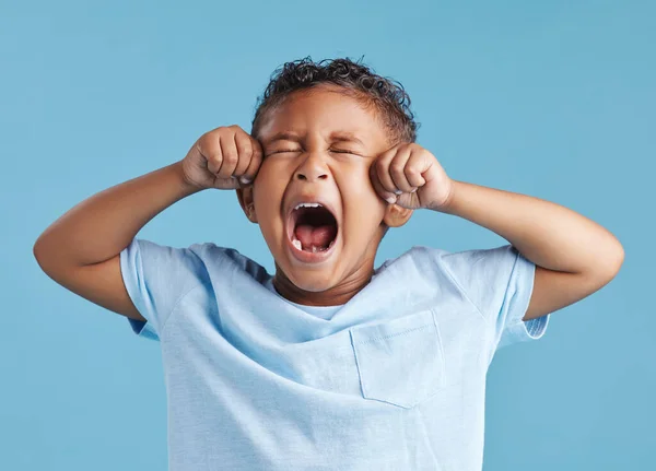 Unhappy little hispanic boy looking upset and crying while rubbing his eyes against a blue studio background. Unhappy preschooler kid bawling his eyes out.
