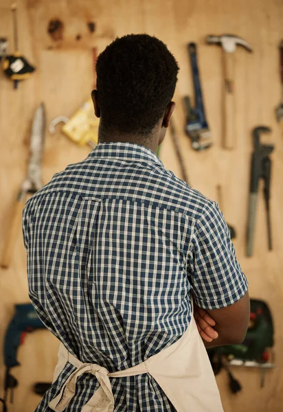 Back of african american carpenter choosing his tools. Designer standing by a wall of tools and equipment. Creative entrepreneur deciding on tools to use. Businessman choosing equipment