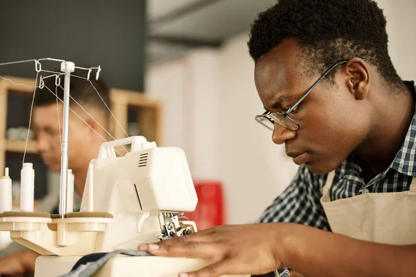 Focused designer sewing fabric. Focused tailor using sewing machine. African American designer sewing a piece of denim. Young businessman working on a sewing machine. Tailor sewing a garment.