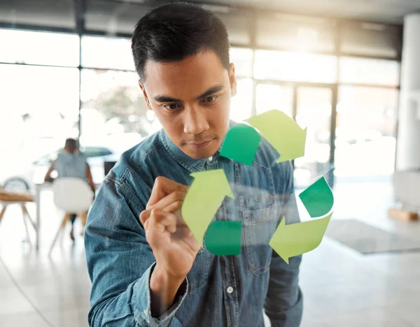 Young focused mixed race businessman drawing a recycle symbol on a glass window in an office at work. One serious hispanic businessperson designing a sign for awareness to recycling.