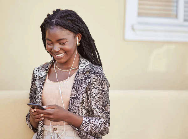 Stylish african american woman with using earphones to listen to music on smartphone or watch funny video online. Smiling young woman using mobile app while walking on city street.