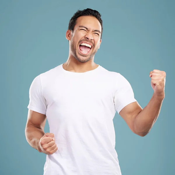 Handsome young mixed race man celebrating victory or success while standing in studio isolated against a blue background. Hispanic male cheering and pumping his fists at success or achievement.