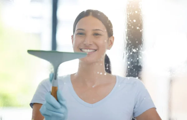 Portrait of a happy mixed race domestic worker using a squeegee on a window. One Hispanic woman enjoying doing chores in her apartment.