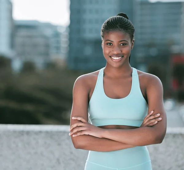 Portrait of one confident young african american woman standing with arms crossed ready for exercise outdoors. Determined female athlete looking happy and motivated for training workout in the city.