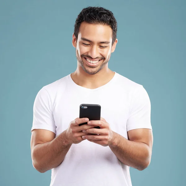 Handsome young mixed race man using his phone while standing in studio isolated against a blue background. Hispanic male sending a text message, using the internet online or browsing social media.