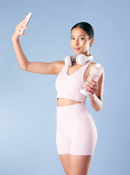 Mixed race fitness woman posing with her phone in studio against a blue background. Young hispanic female athlete taking selfie pictures with her smartphone to track her personal fitness growth.