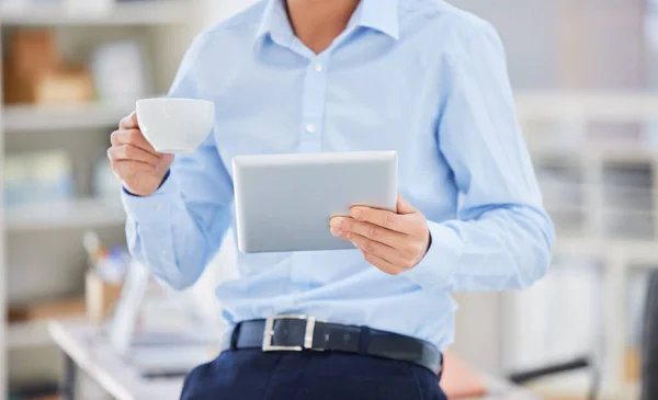 Closeup of on businessman browsing on a digital tablet while drinking coffee from a teacup during a break in an office. Entrepreneur planning online while searching for ideas and inspiration on his
