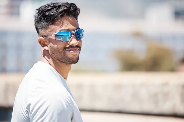 Fit young male athlete wearing sports sunglasses and smiling while looking at the camera. Mixed race sportsman out for a run or jog in the city.