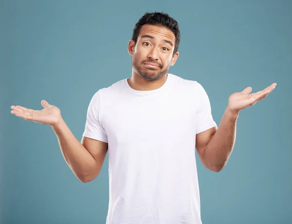 Handsome young mixed race man shrugging his shoulders while standing in studio isolated against a blue background. Confused hispanic male looking lost or clueless and making a, So what gesture.