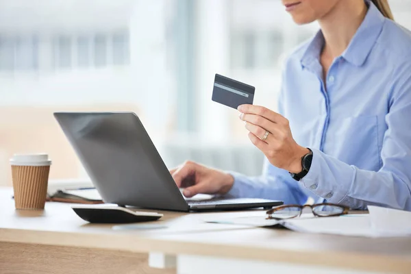 Closeup of caucasian woman reading her credit card while using her phone and laptop to shop online while sitting in the office at work. Shopping has never been simpler or more convenient.