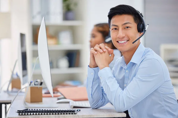 Portrait of one happy young asian male call centre telemarketing agent talking on a headset while working on a computer in an office. Confident and friendly businessman consultant operating a helpdesk