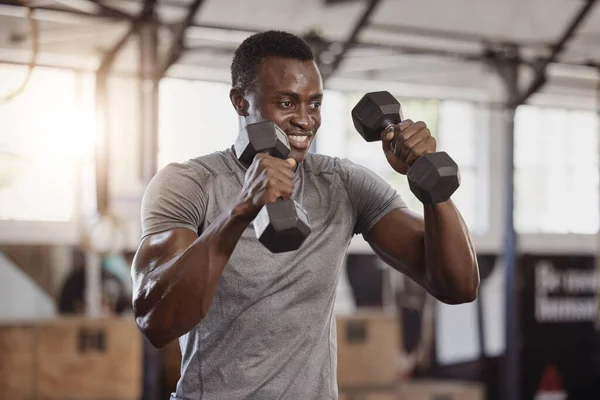 Smiling young african american athlete lifting dumbbells during arm workout in gym. Strong, fit, active happy black man training with weights in health and sport club. Weightlifting exercise routine.