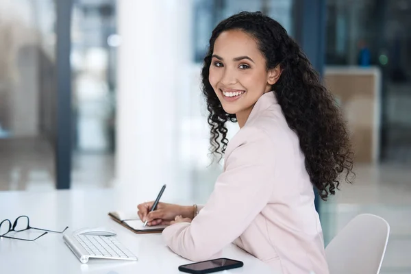 Portrait of smiling mixed race business woman sitting at her desk. Ambitious professional writing in journal while sitting in her office. Entrepreneur planning her business day.