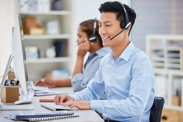Happy young asian male call centre telemarketing agent talking on a headset while working on a computer in an office. Confident and friendly businessman consultant operating a helpdesk for customer