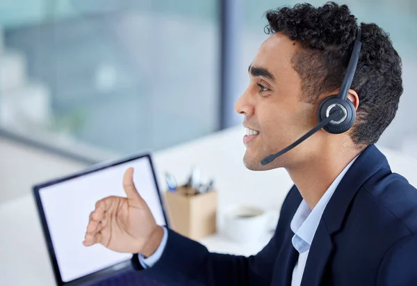 One happy young mixed race male call centre telemarketing agent talking on a headset while working on a laptop with blank screen in an office. Confident and friendly hispanic businessman consultant