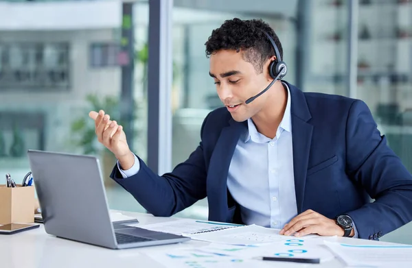 One young mixed race call centre agent talking on a headset while reading document notes and using laptop for video call in an office. Hispanic salesman and marketing rep consultant advising while