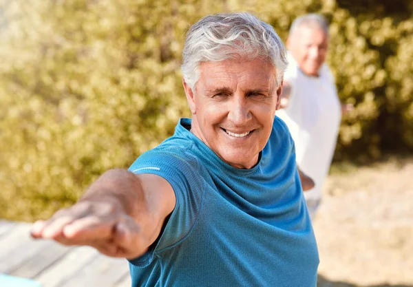 Happy energetic senior man standing in warrior pose while practicing yoga during fitness class in nature on a sunny day. Seniors living a healthy lifestyle and staying active.