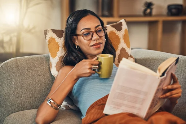Beautiful mixed race woman reading book and drinking coffee in living room at home. Hispanic lying down on lounge sofa alone and enjoying novel. Feeling relaxed on weekend with fiction story and tea.
