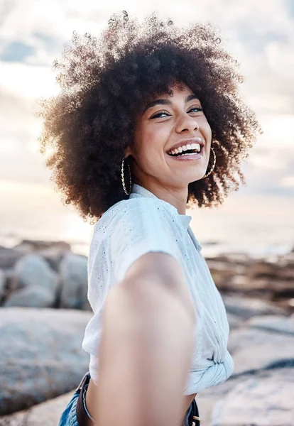 A young mixed race woman smiling on the beach while reaching out her hand. Happy hispanic female with a cool afro hairstyle outdoors.