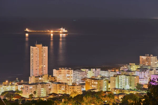 View of skyscraper and city buildings lighting up the dark sky at night, alongside the ocean horizon. Beautiful scenic urban landscape in the evening with a seascape background with a ship at sea