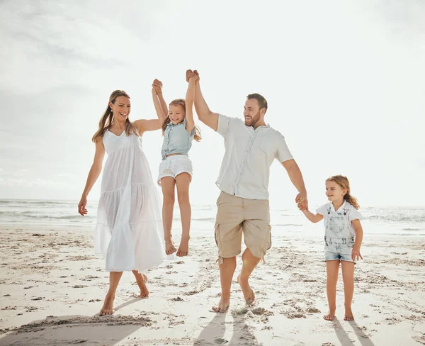 Happy caucasian family of four having fun while enjoying a summer vacation together at the beach shore. Loving parents swinging their cute kids while walking on sand. Bonding and sharing quality time.