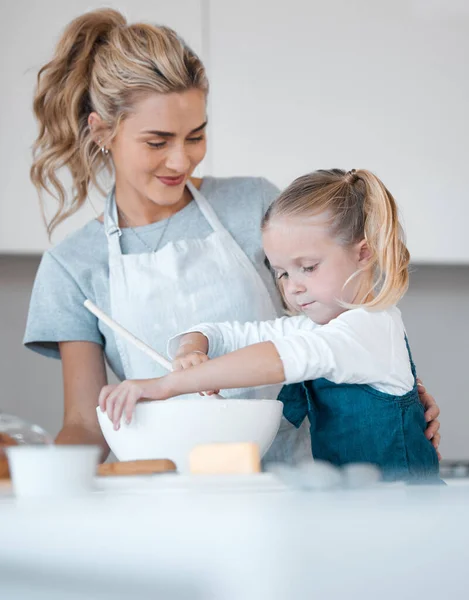 Happy mother helping her child bake. Woman looking at her daughter bake. Caucasian little girl mixing a bowl of batter. Proud mother watching her daughter bake. Small girl enjoying baking.