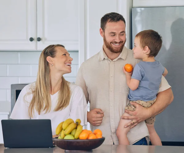 Happy young caucasian family having lunch time with fresh fruit in bright kitchen. Beautiful young mother and father feeding their cute son an orange while using a laptop at home.