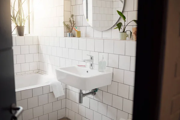 A clean and tidy bathroom inside a house. A basin, bath, with ceramic tiling and a round mirror hanging on the wall. A clean, hygienic, bright room.