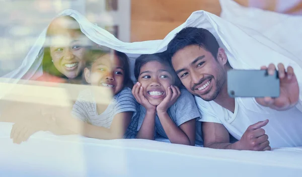 Happy parents with little children lying on bed at home under blanket and taking selfies on cellphone. Smiling mixed race girls bonding and enjoying free time with their mother and father.