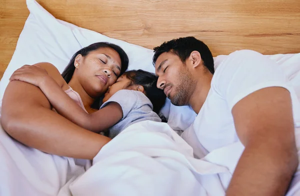 Mixed race family of three asleep in bed. Above mother, father and daughter resting on a white bed in the bedroom at home. Tired parents lying in bed sleeping with their daughter in the middle.