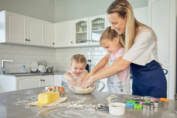 Caucasian caring mother and little daughters baking together in a kitchen at home. Mom teaching girls how to make dough in a messy kitchen. Sisters learning how to bake with their mom.