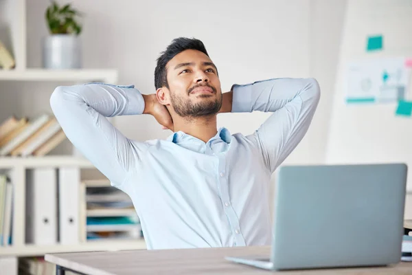 Young mixed race businessman resting with his arms behind his back working on a laptop in an office at work. Hispanic man relaxing while thinking. Male boss enjoying success alone at work.