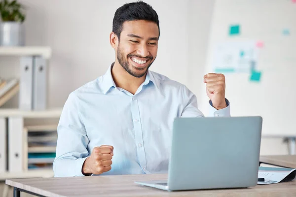 Young happy and excited mixed race businessman cheering with his fist while working on a laptop alone in an office at work. One hispanic male boss smiling while celebrating success.