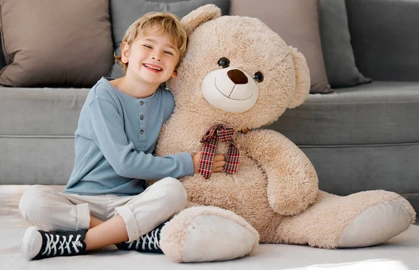 Portrait of one happy little caucasian boy smiling while hugging a big and cosy fluffy teddy bear on the floor in the lounge at home. Adorable kid relaxing and playing with soft stuffed toy alone.