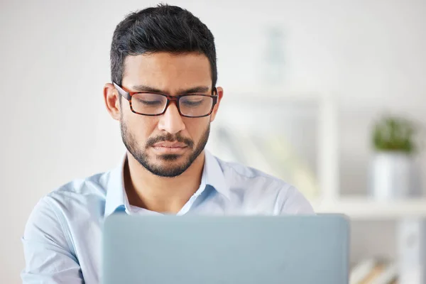 Young focused mixed race businessman working alone on a laptop in an office at work. Serious hispanic male boss wearing glasses while reading an email on a laptop.