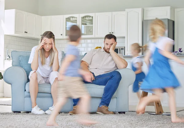 Stressed parents sitting on couch with children running around.Excited children playing around upset parents. Frustrated couple children running around. Unhappy parents upset at kids. Children playin.